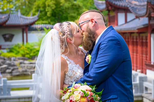 Romantischer Moment: Hochzeit im Japanischen Garten zu Erfurt Nahaufnahme eines Brautpaares, das sich in einem japanischen Garten vor einem Gebäude und einem Teich küsst. Der Fokus liegt auf den Gesichtern und dem bunten Brautstrauß.